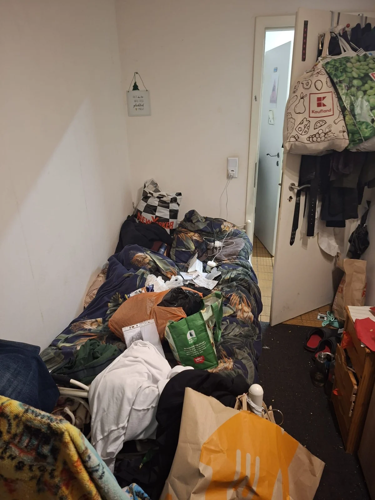 Ein kleiner Raum mit einem durcheinandergebrachten Bett, Kleidung und Einkaufstaschen darauf. Die Tür steht offen.