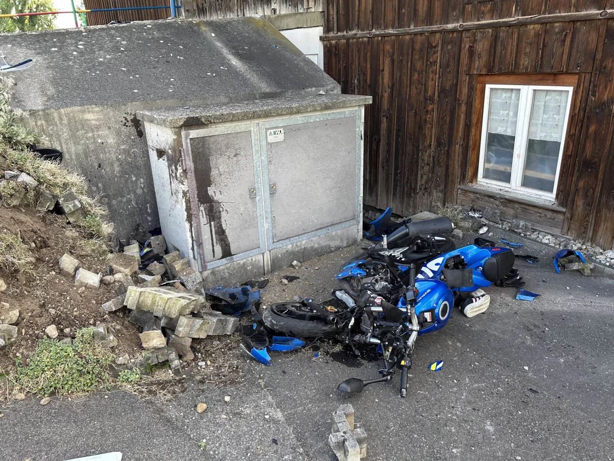 Ein blaues Motorrad liegt stark beschädigt vor einem Gebäude, umgeben von Betonblöcken und Trümmern. Im Hintergrund ist eine Holzwand und ein Fenster zu sehen.