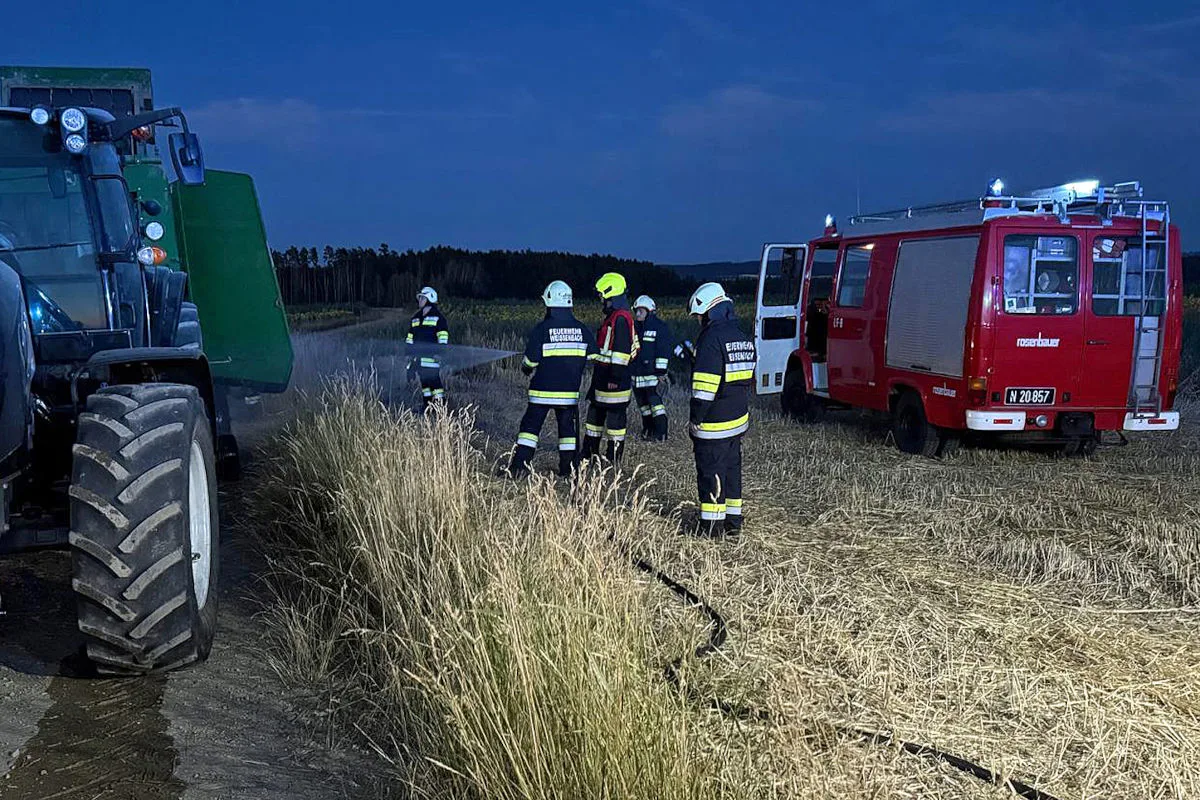 Mehrere Feuerwehrleute in Uniform stehen auf einem Feld neben einem Traktor und einem Feuerwehrauto bei Nachtbeleuchtung.