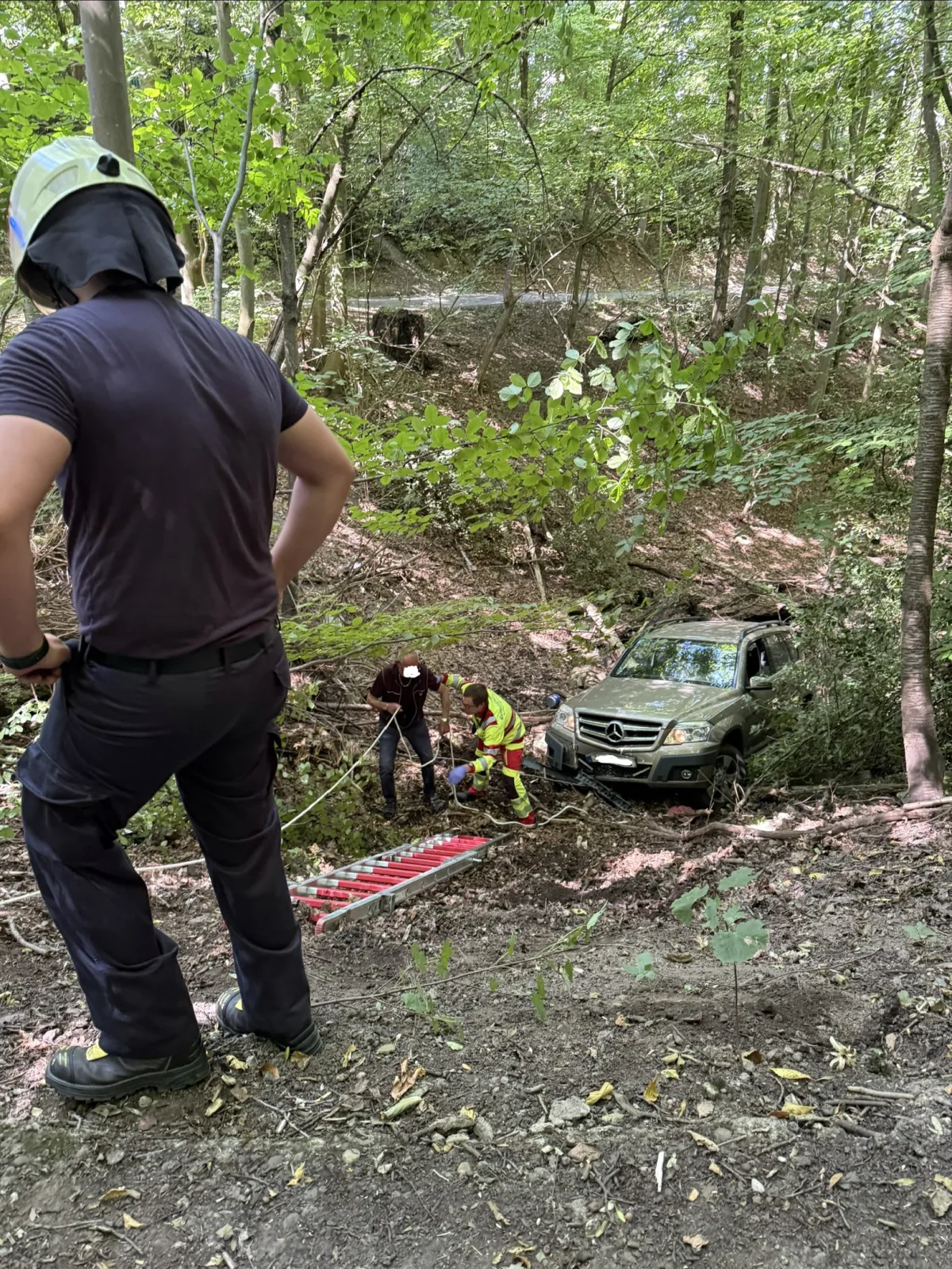 Ein SUV ist im Wald von einem Abhang gerutscht. Mehrere Personen sind bei der Bergung beschäftigt, einer trägt Schutzkleidung.