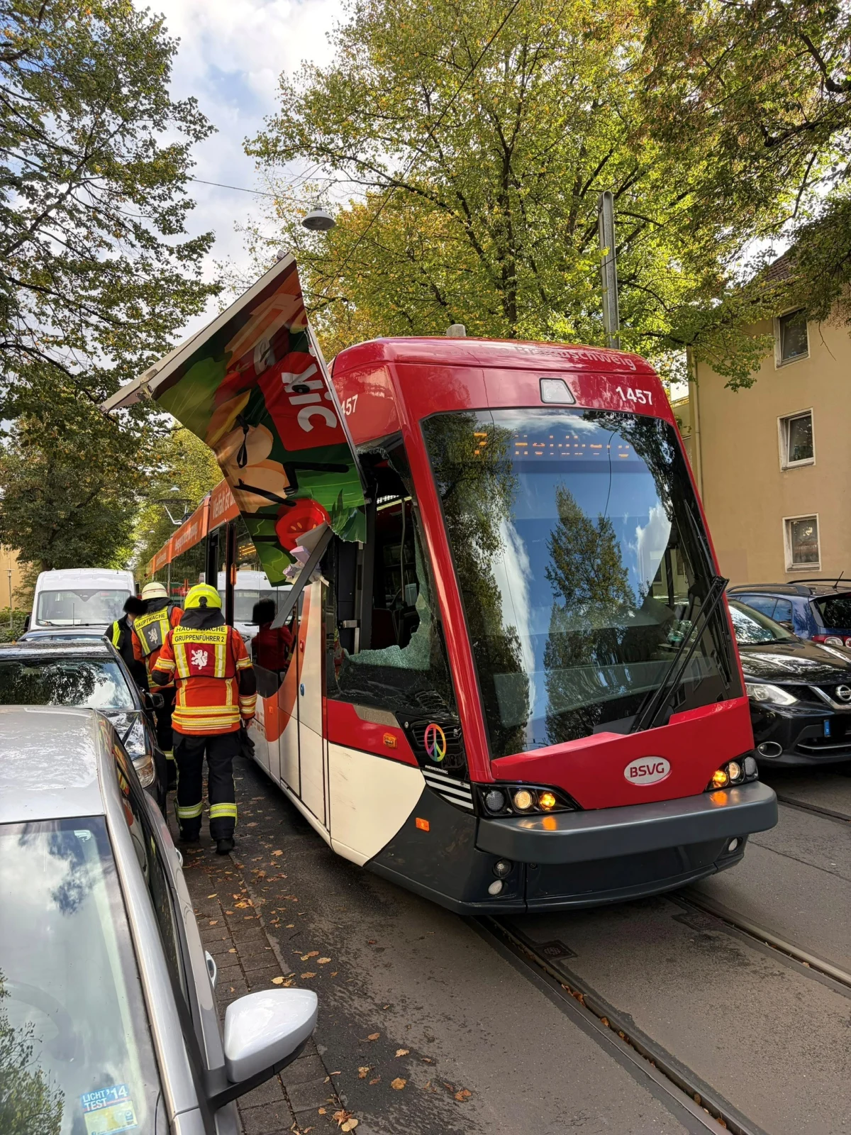 Eine rote Straßenbahn mit beschädigtem Dach steht auf einer Straße. Zwei Feuerwehrleute sind neben der Straßenbahn zu sehen.