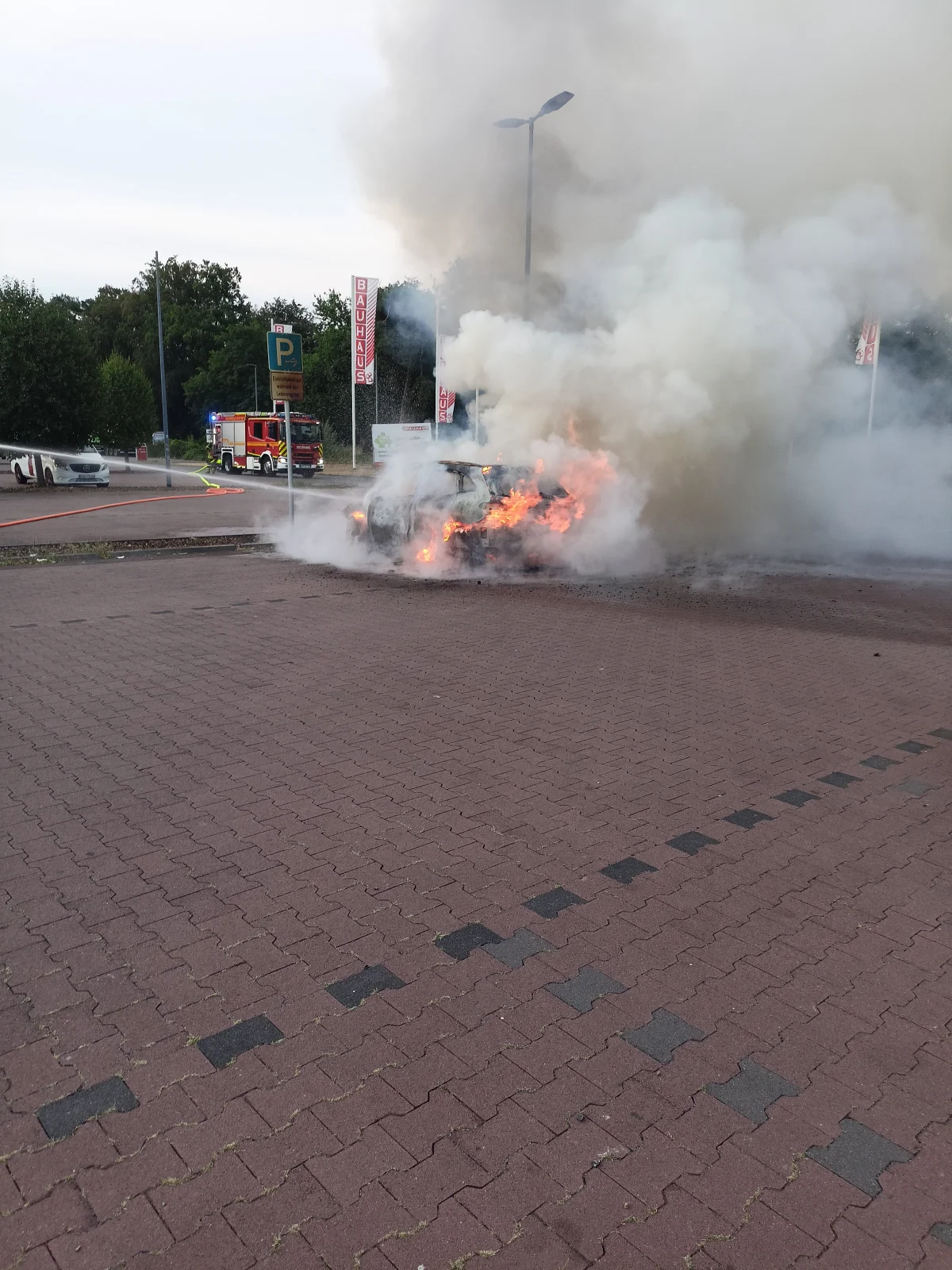 Auf einem Parkplatz brennt ein Fahrzeug, aus dem dichter Rauch aufsteigt. Im Hintergrund ist ein Feuerwehrauto zu sehen.