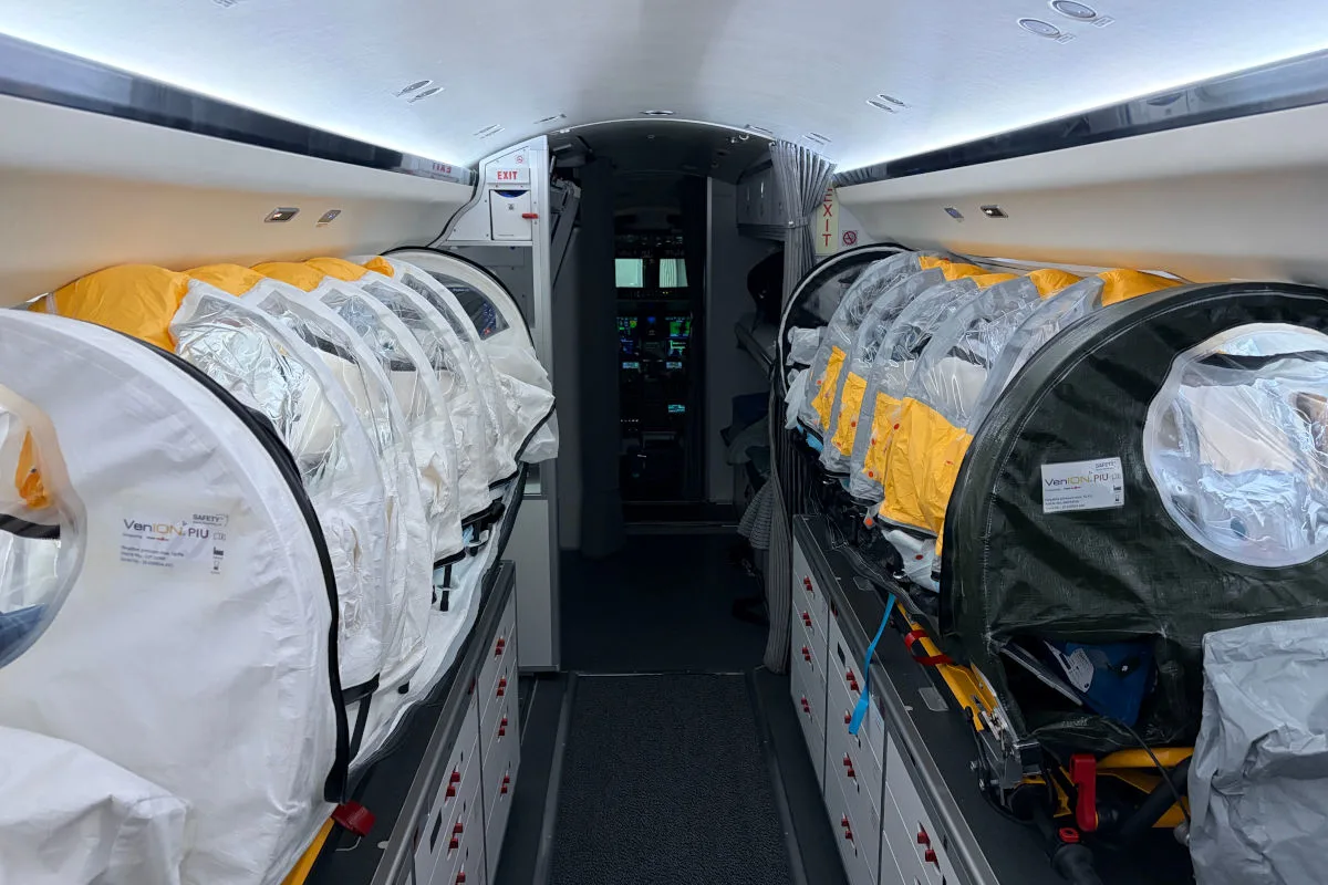 Im Bild ist das Innere eines medizinisch ausgestatteten Flugzeugs zu sehen, das mehrere Isoliertragen für den Patiententransport enthält. Die Tragen sind in einem schmalen Gang mit Blick auf das Cockpit angeordnet.