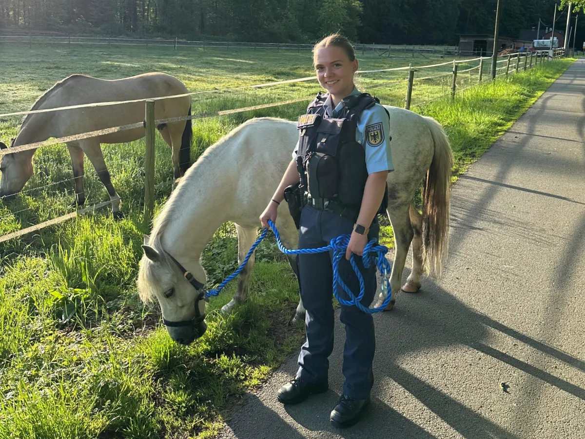 Eine uniformierte Polizistin hält ein weißes Pony, das an einer blauen Leine geführt wird, auf einem asphaltierten Weg neben einer grasbewachsenen Wiese, wo ein weiteres Pferd zu sehen ist.