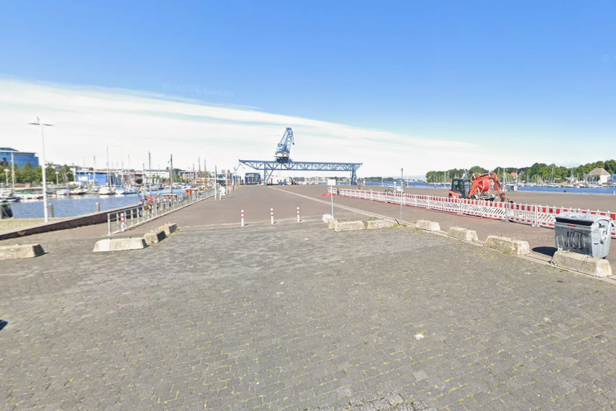 Der Hafen zeigt einen großen Kran in der Mitte und Absperrungen auf der Straße. Der Himmel ist klar und blau, mit vereinzelten Wolken.
