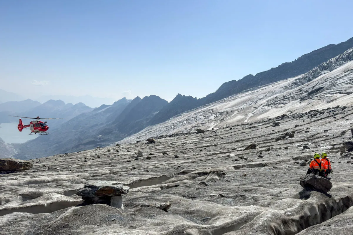 Ein Rettungshubschrauber fliegt über einen weitläufigen Gletscher in einer Berglandschaft. Zwei Personen in leuchtenden Jacken stehen nahe einer Felskante.