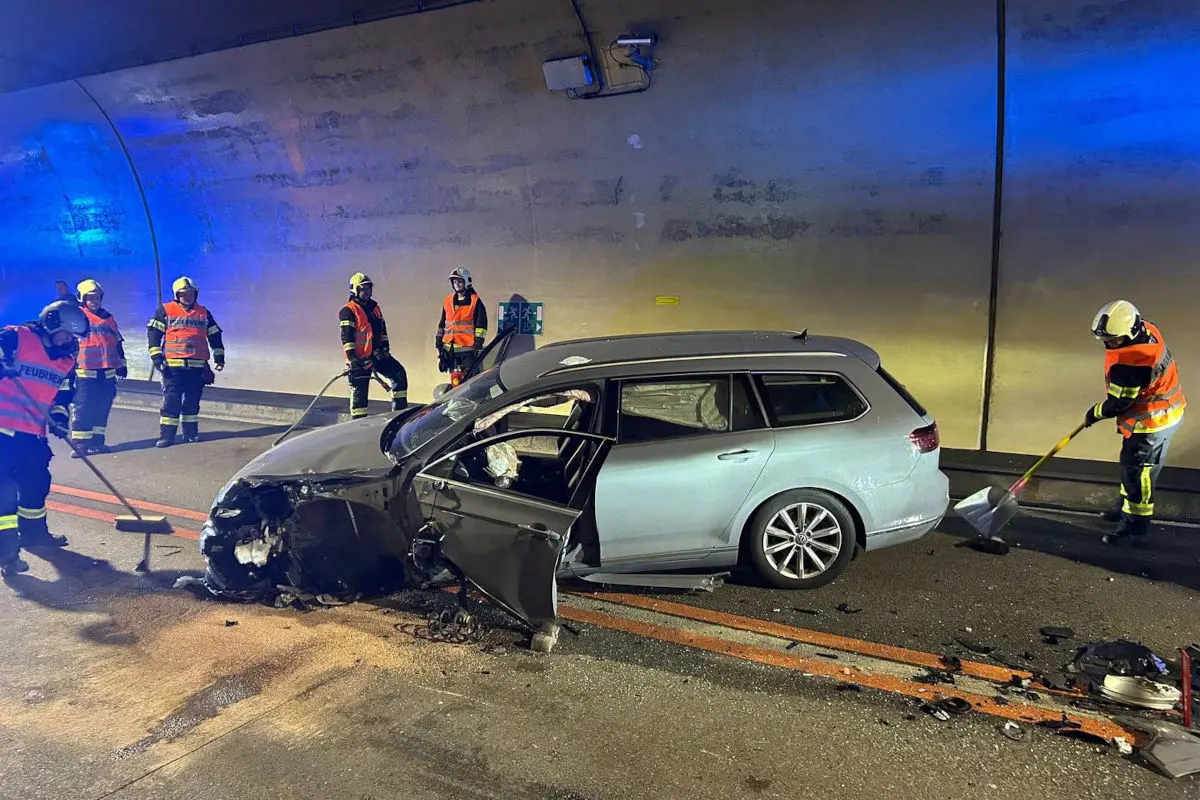 In einem Tunnel steht ein stark beschädigtes Auto, umgeben von mehreren Feuerwehrleuten in orangefarbenen Warnwesten. Diese sichern die Unfallstelle ab und beseitigen Trümmer von der Straße.