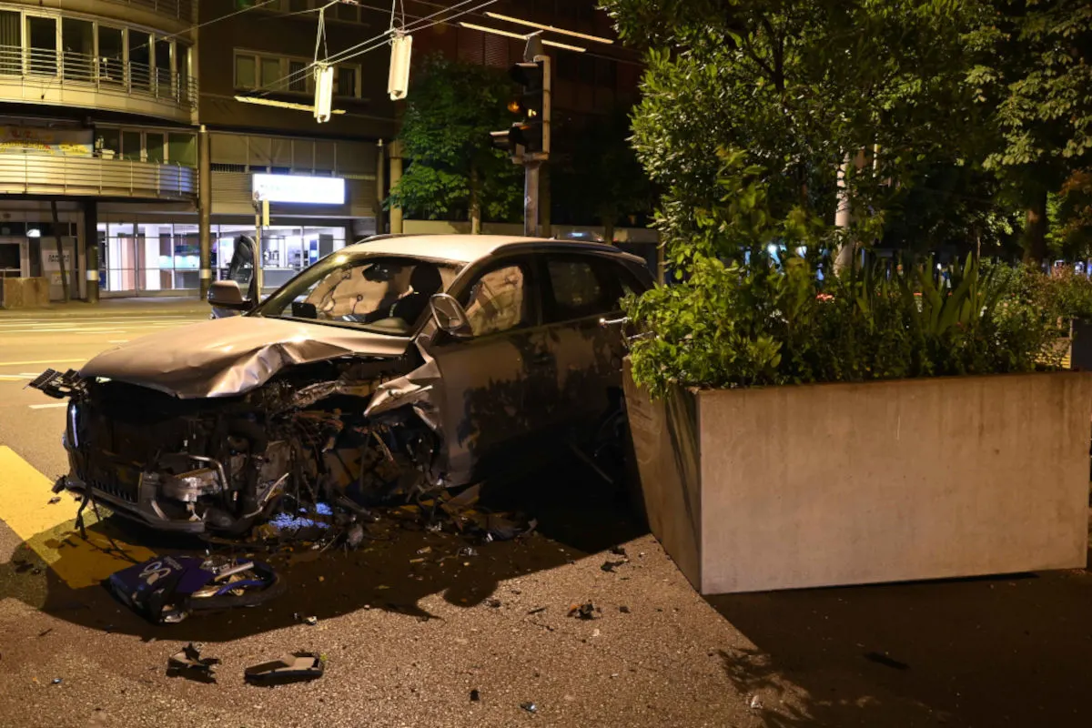 Das Bild zeigt ein Auto mit stark beschädigter Frontpartie, das gegen einen großen Betonpflanzkübel gefahren ist. Es ist Nacht und die Szene ist in einer städtischen Umgebung aufgenommen.