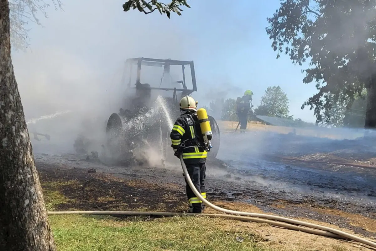 Ein Feuerwehrmann spritzt Wasser auf ein brennendes Fahrzeug, während Rauch aufsteigt. Ein weiterer Feuerwehrmann ist im Hintergrund sichtbar.