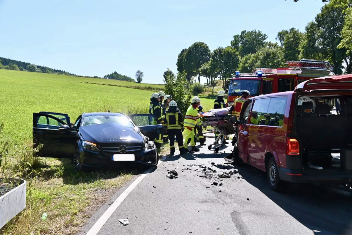 Feuerwehrleute stehen neben einem beschädigten schwarzen Mercedes auf einer Landstraße. Ein Feuerwehrfahrzeug und ein Einsatzwagen sind ebenfalls am Ort.