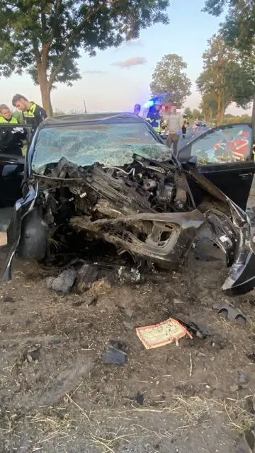 Ein stark zerstörtes Fahrzeug steht mit geöffneten Türen auf einer ländlichen Straße, umgeben von Bäumen. Polizei und Rettungskräfte sind im Hintergrund zu sehen.