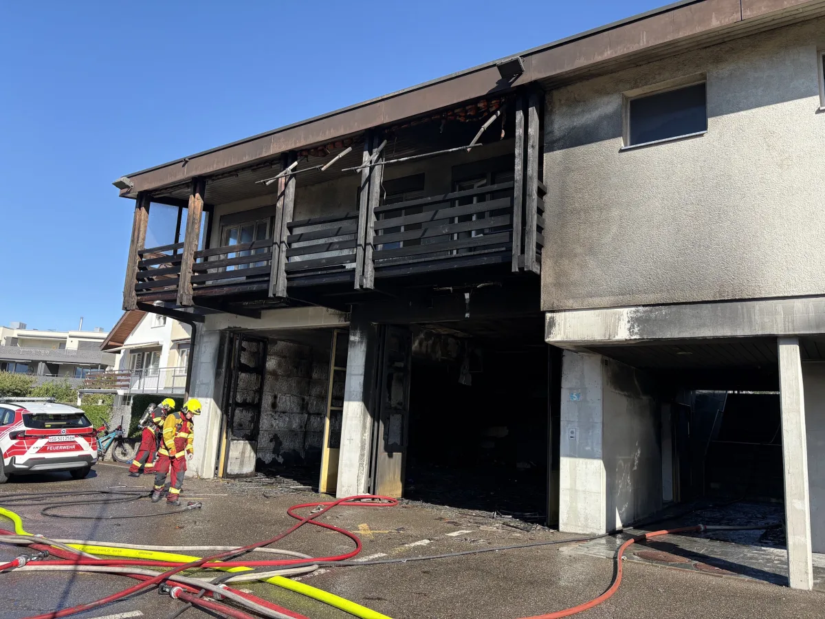 Mehrere Feuerwehrleute in voller Montur stehen vor einem Wohnhaus mit sichtbaren Brandschäden an einem Balkon. Ein Feuerwehrfahrzeug ist im Vordergrund erkennbar.