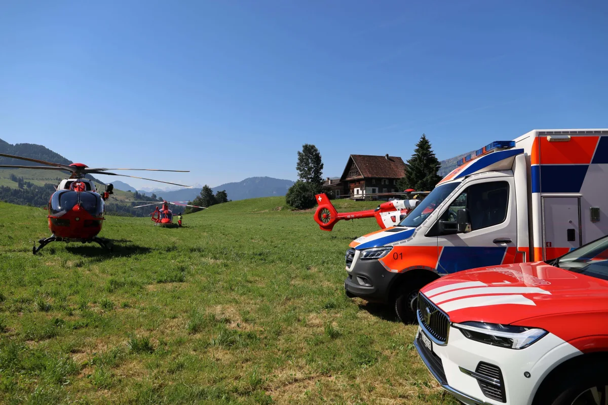 Zwei rot-weiße Rettungshubschrauber stehen auf einer grünen Wiese. Im Hintergrund sind Notfallfahrzeuge und ein Bauernhaus sichtbar.