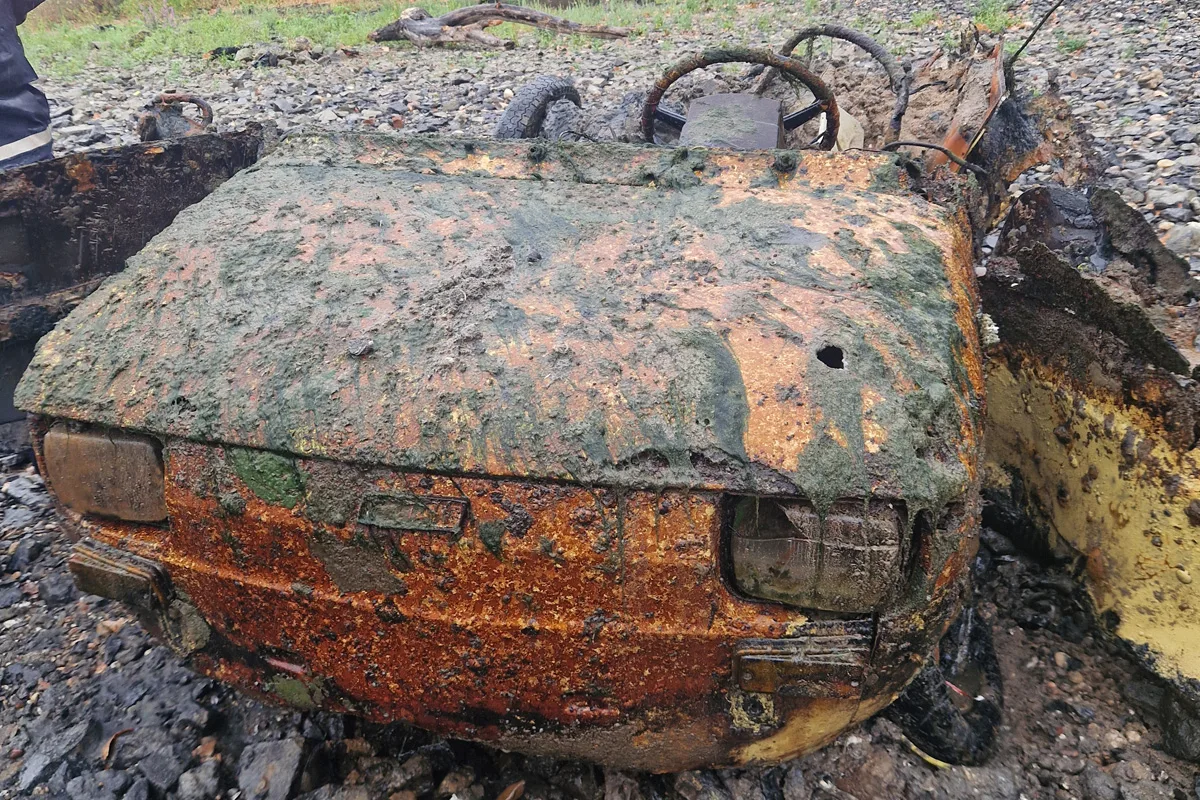 Ein stark verrostetes Autowrack mit sichtbarem Lenkrad liegt auf einer steinigen Fläche. Das Fahrzeug ist mit Schmutz und Rost bedeckt.