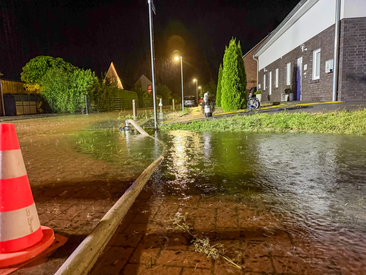 Eine Nachtaufnahme zeigt eine überflutete Straße in einem Wohngebiet, wo eine Pumpe Wasser abführt. Im Vordergrund erkennbar eine Absperrung und Wasser auf der Straße.