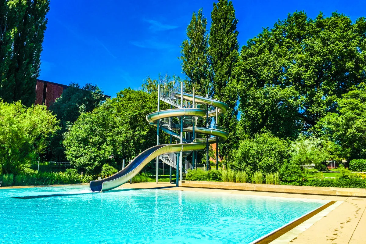 Eine spiralförmige Rutsche mündet in einen leeren Außenpool. Im Hintergrund sind Bäume und ein klarer blauer Himmel zu sehen.