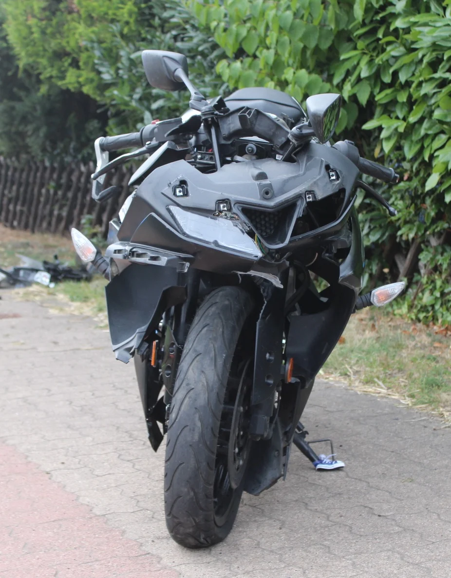 Ein schwarzes Motorrad steht beschädigt auf einem Gehweg, umgeben von Pflanzen und einem Holzzaun.