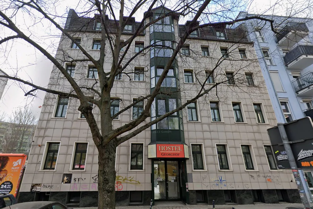 Ein graues Gebäude mit dem Schild 'HOSTEL Georgshof' über dem Eingang. Vor dem Gebäude steht ein kahler Baum.