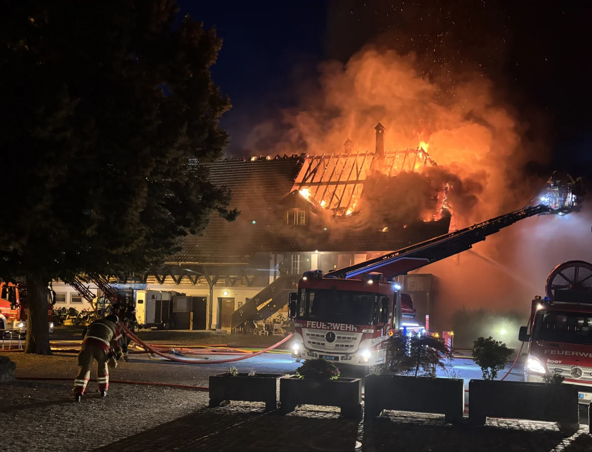 Ein Feuerwehreinsatz bei Nacht: Das Dach eines Gebäudes steht in Flammen, mehrere Feuerwehrfahrzeuge sind vor Ort. Feuerwehrleute arbeiten auf einer Leitern, um das Feuer zu löschen.