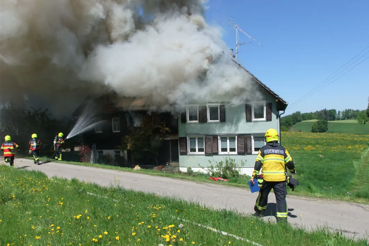 Feuerwehrleute mit gelben Helmen und Schutzanzügen löschen ein Hausbrand unter starkem Rauch. Es ist ein sonniger Tag.