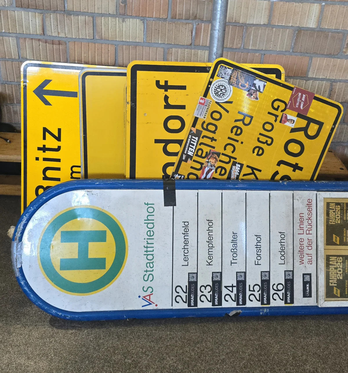 Mehrere gestapelte Verkehrsschilder und ein Busfahrplan sind an einer Backsteinwand angelehnt. Die Schilder sind gelb mit schwarzer Schrift.