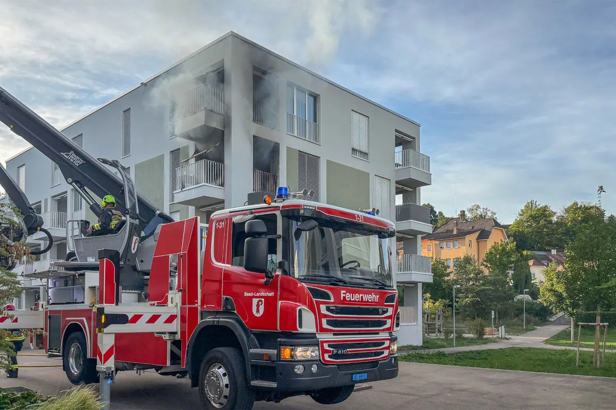 Ein Feuerwehrfahrzeug mit Drehleiter ist vor einem modernen Wohnhaus im Einsatz zu sehen. Rauch steigt aus einem der oberen Stockwerke.