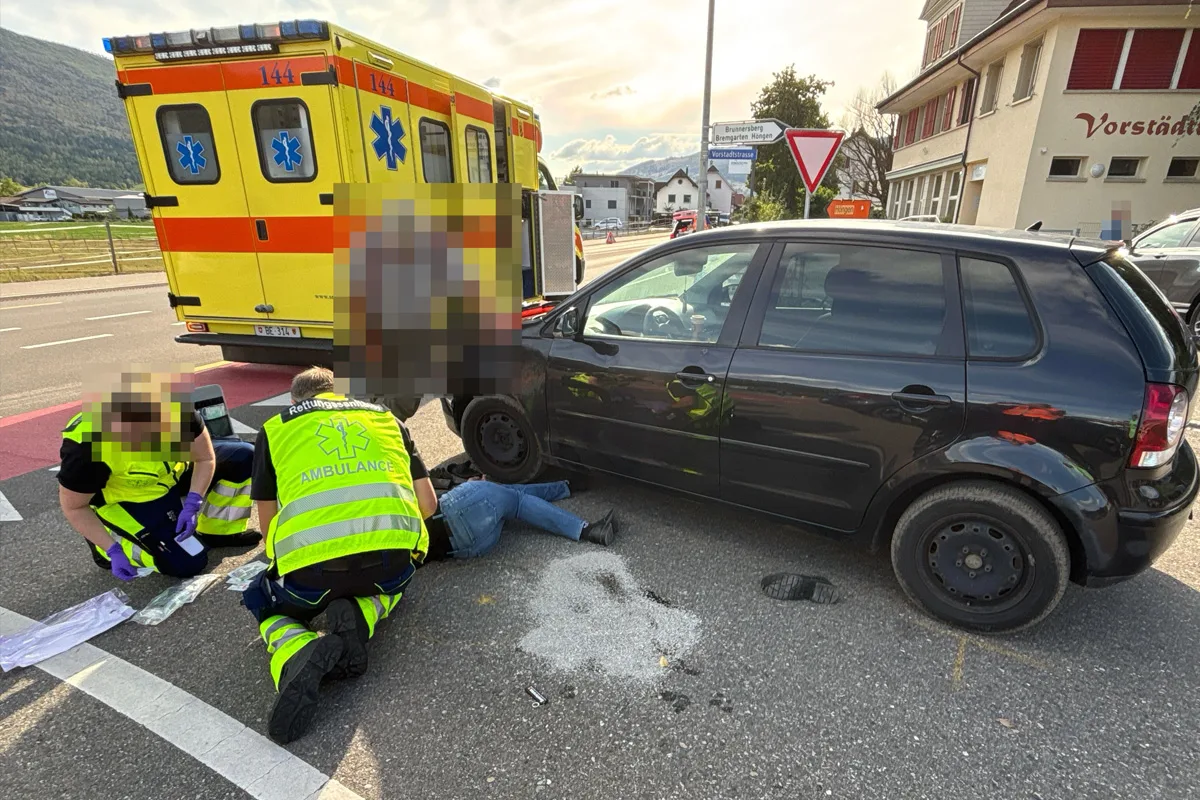 Sanitäter kümmern sich um eine liegende Person neben einem schwarzen Auto und einem gelben Rettungswagen auf einer Kreuzung bei Tageslicht.