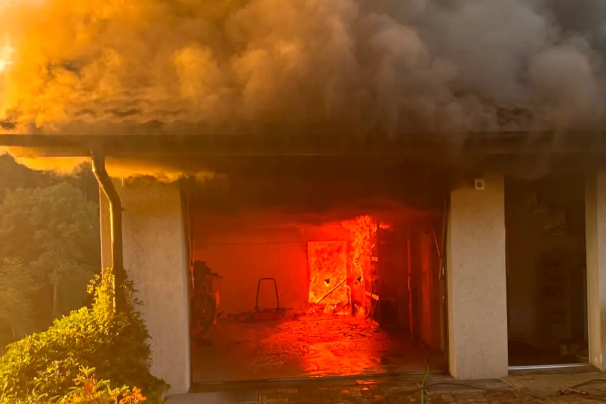 In einer Garage brennt ein intensives Feuer, das helle Flammen und dichten Rauch erzeugt. Schwarzer Rauch dringt aus der Öffnung, während das Innere rot erleuchtet ist.