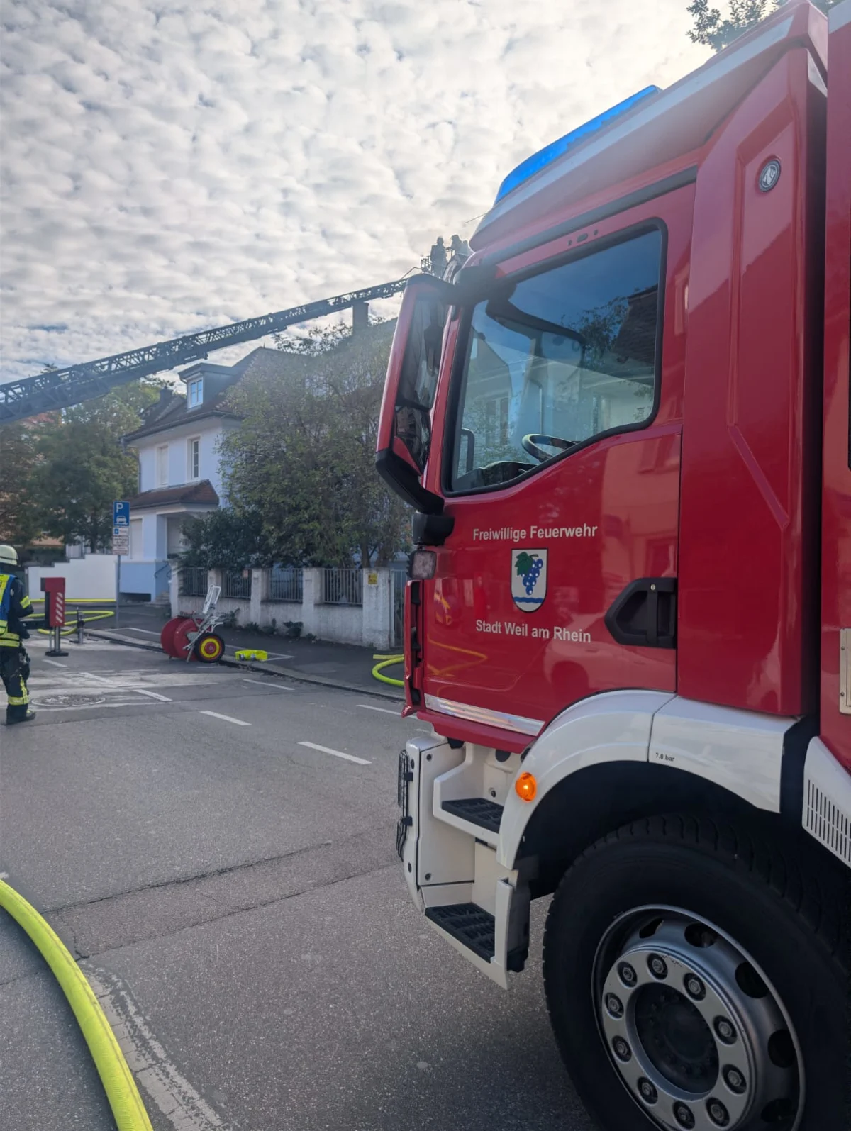 Ein Feuerwehrfahrzeug der Freiwilligen Feuerwehr Weil am Rhein steht auf einer Straße. Eine Drehleiter ist ausgefahren, und Feuerwehrleute sind im Einsatz.