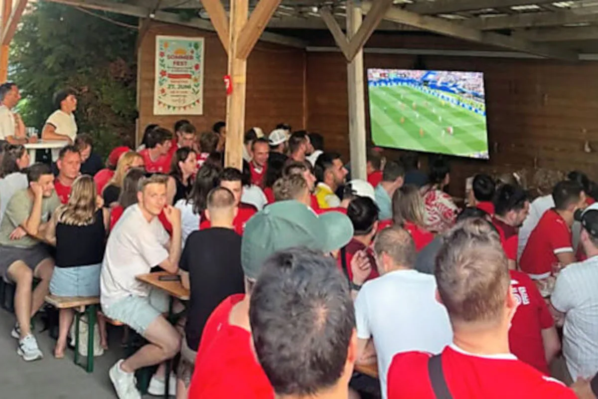 Eine große Gruppe von Menschen sitzt in einem überdachten Biergarten und schaut ein Fußballspiel auf einem großen Bildschirm. Viele der Personen tragen rote Kleidung, vermutlich Fanutensilien.