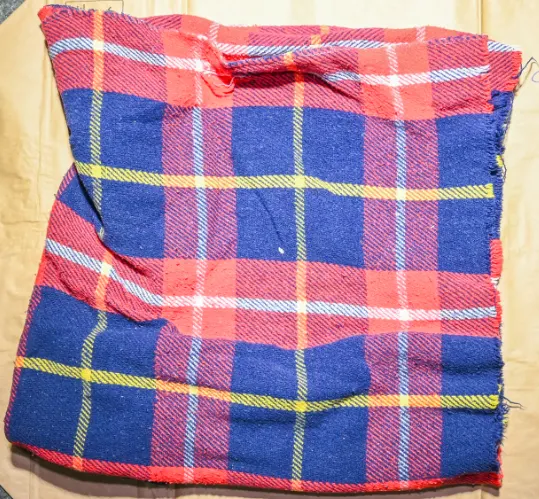 Ein karierter Schal in Blau und Rot mit gelben Streifen liegt auf einem hellen Untergrund. Der Stoff wirkt leicht verknittert und hat kleine Risse.