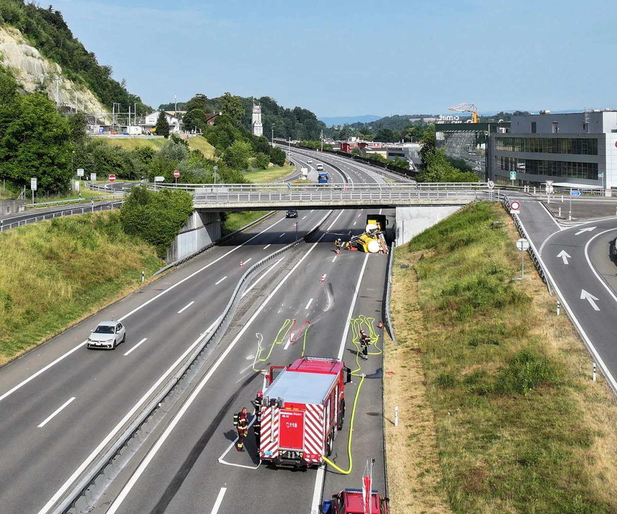 Ein Feuerwehrauto steht auf einer Autobahn, während ein umgekippter Lkw dahinter sichtbar ist. Feuerwehrleute sind vor Ort im Einsatz.