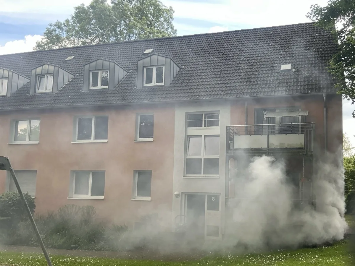 Ein mehrstöckiges Wohnhaus mit sichtbarem Rauch, der von einem Balkon oder Fenster aufsteigt. Das Bild zeigt eine typische städtische Umgebung bei Tageslicht.
