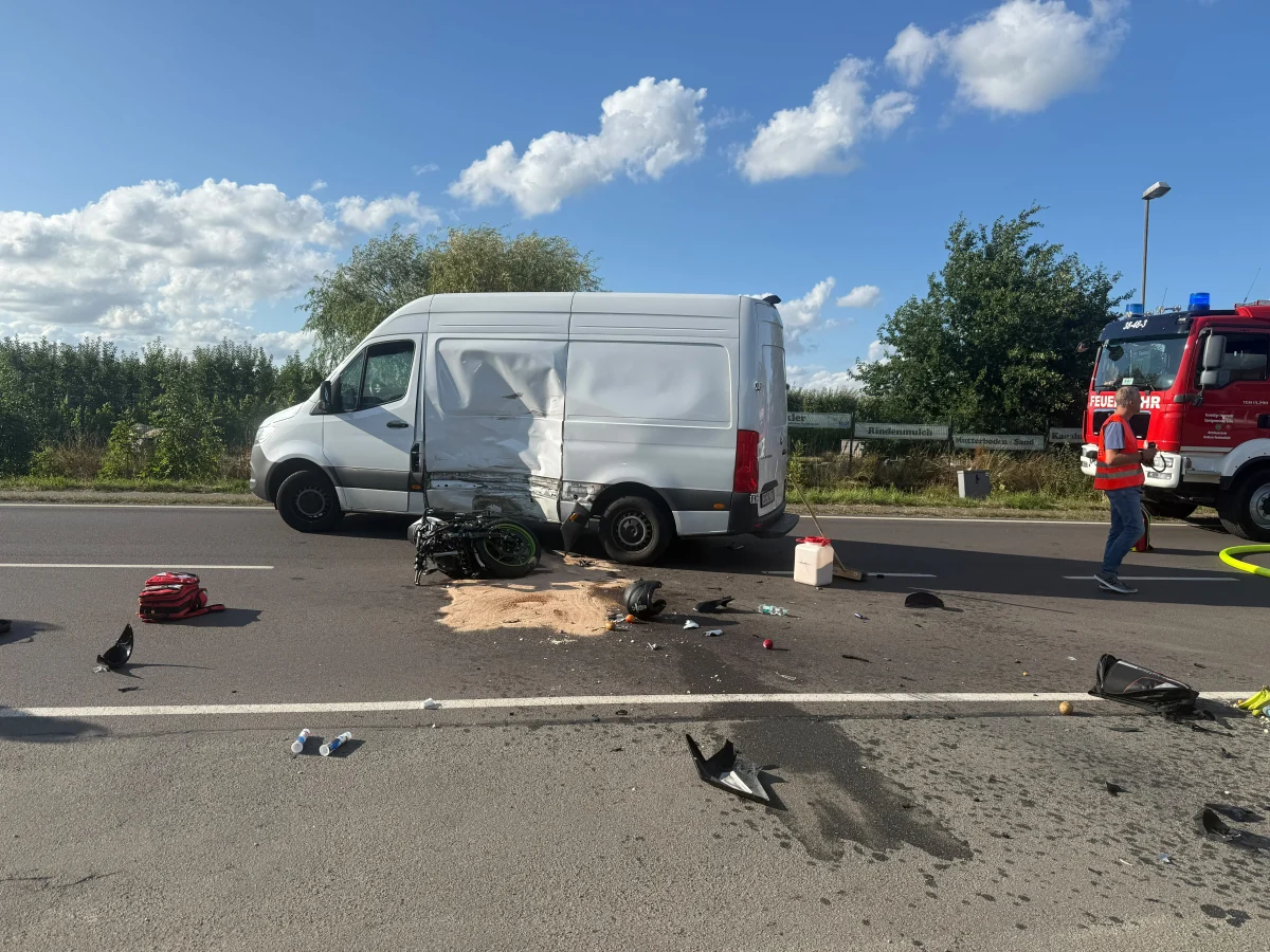 Ein weißer Transporter mit schweren Beschädigungen steht auf der Straße. Daneben liegt ein gestürztes Motorrad und Trümmer sind verteilt. Ein Feuerwehrwagen steht im Hintergrund, ein Mann mit Warnweste ist in der Nähe.