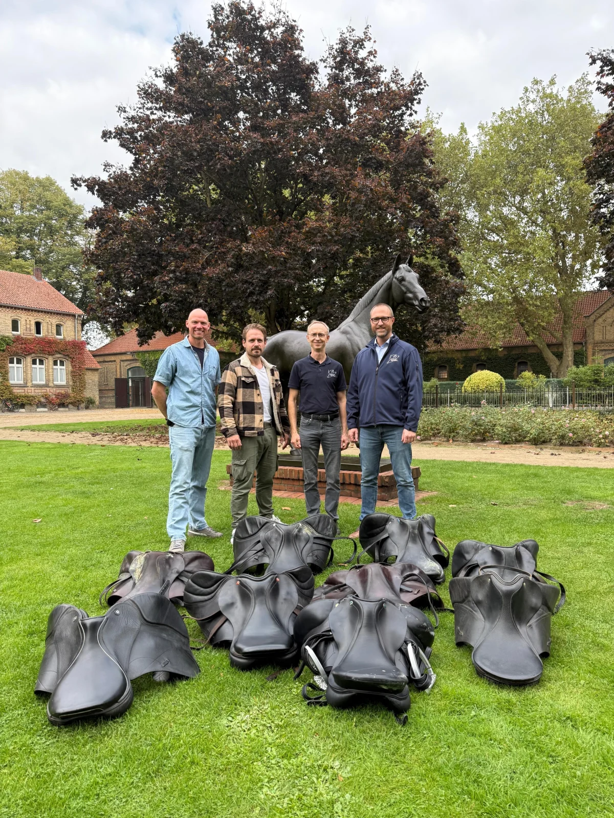Vier Männer stehen auf einer Rasenfläche vor einer Pferdestatue, mehrere schwarze Sättel liegen vor ihnen auf dem Gras.