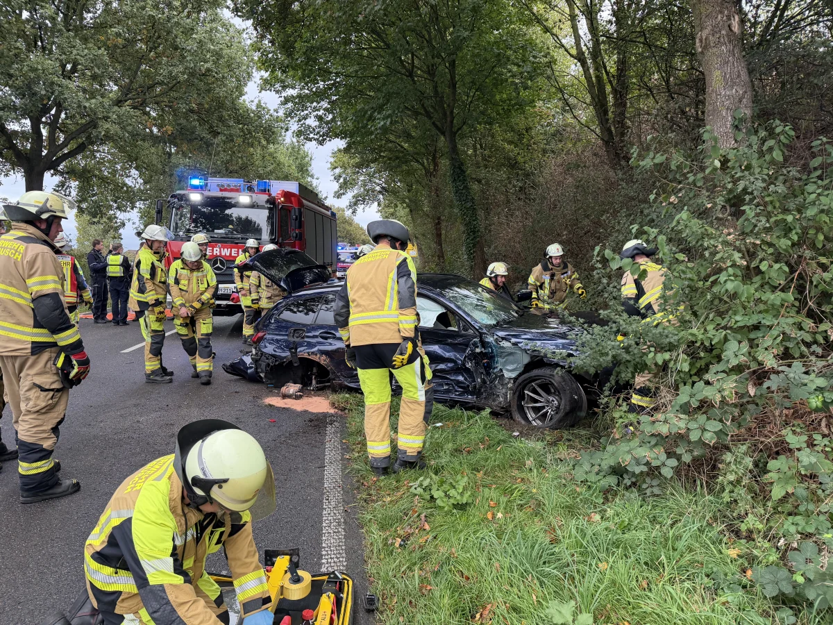 Ein schwer beschädigtes Auto steht nach einem Unfall neben einem Baum. Feuerwehrleute in Einsatzkleidung arbeiten am Unfallort.