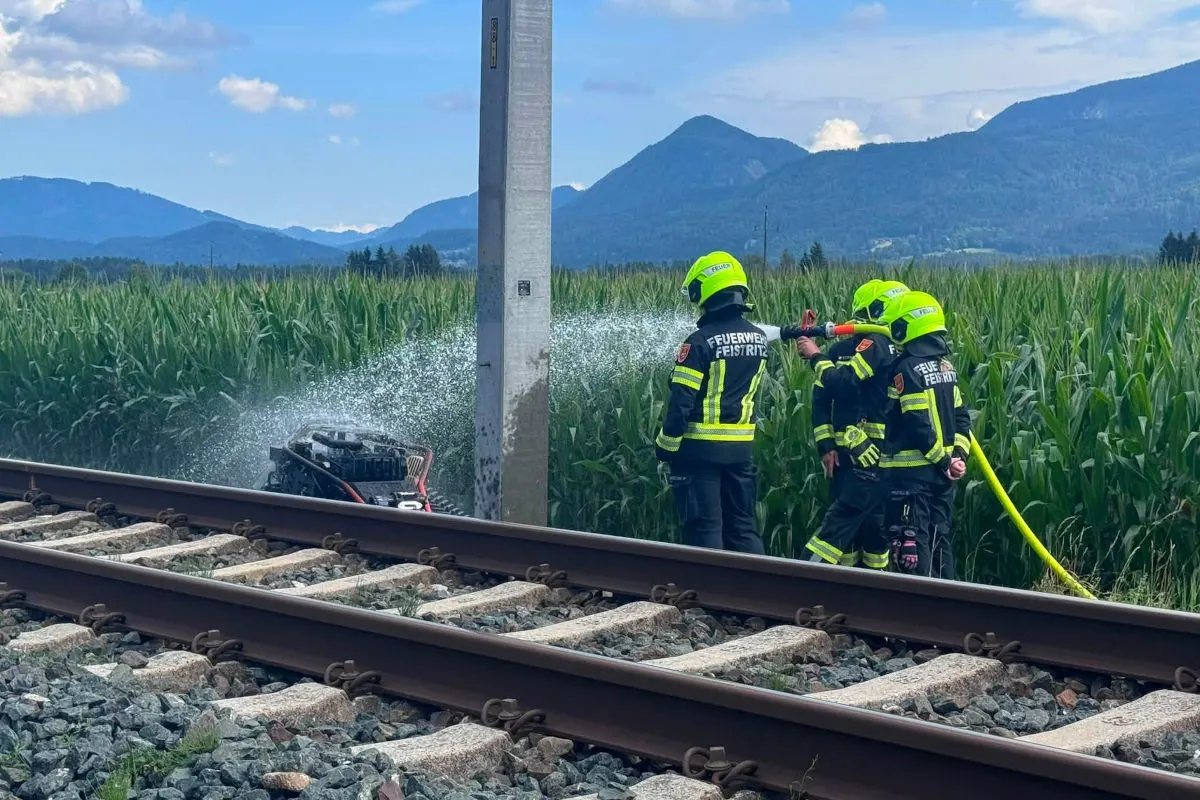Vier Feuerwehrleute bekämpfen ein Feuer an einem Bahngleis mit einem Wasserschlauch. Im Hintergrund sind Berge zu sehen.