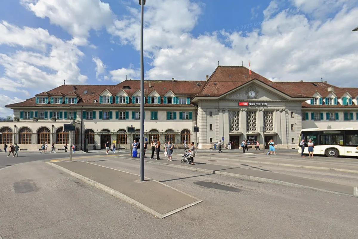Ein großes historisches Bahnhofsgebäude mit rotem Dach, umgeben von Passanten und einem vorbeifahrenden Bus. Der Himmel ist klar und sonnig.