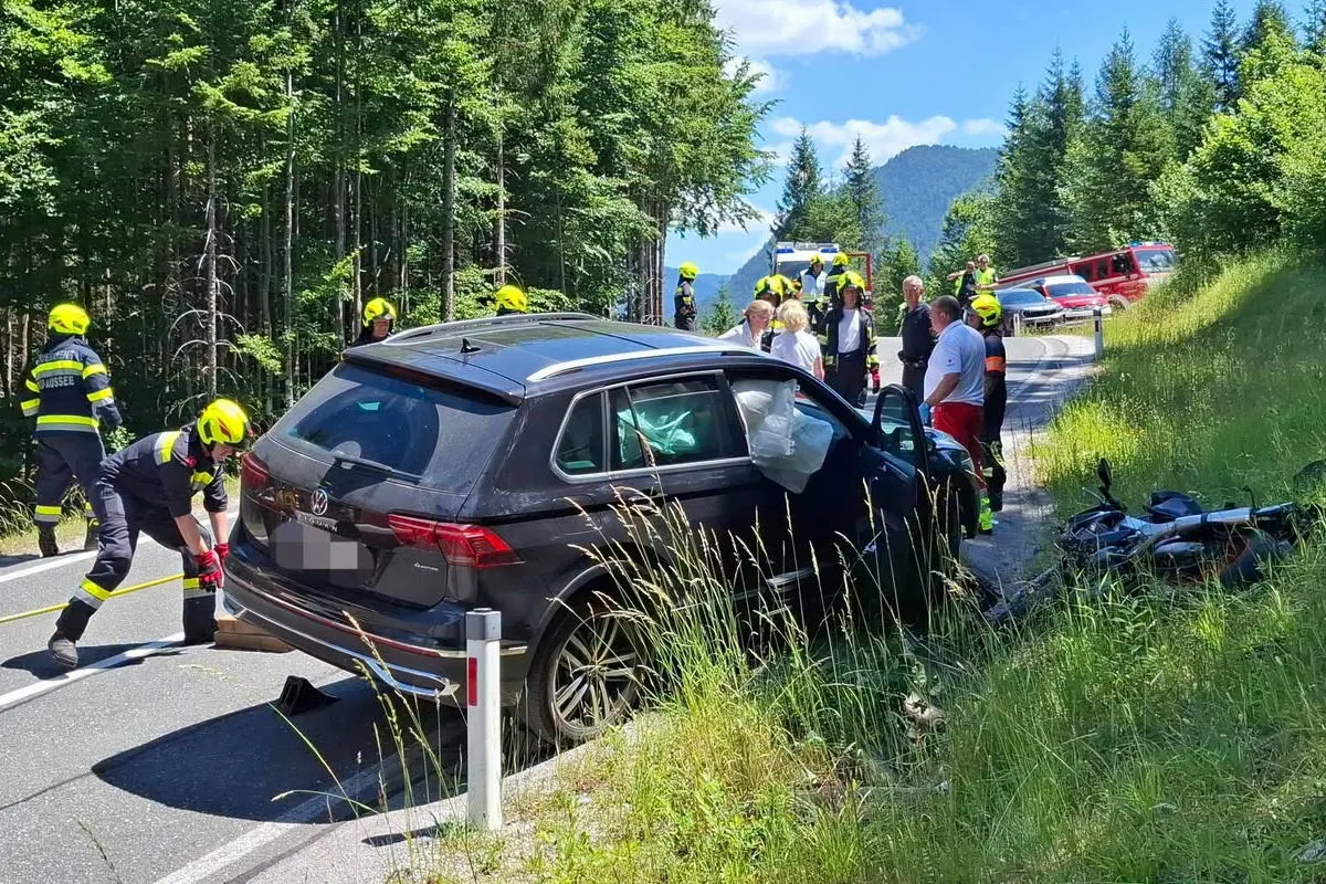 Ein schwarzes SUV steht beschädigt auf der Straße neben einem Motorrad. Feuerwehrleute und Zivilisten sind an der Unfallstelle aktiv.