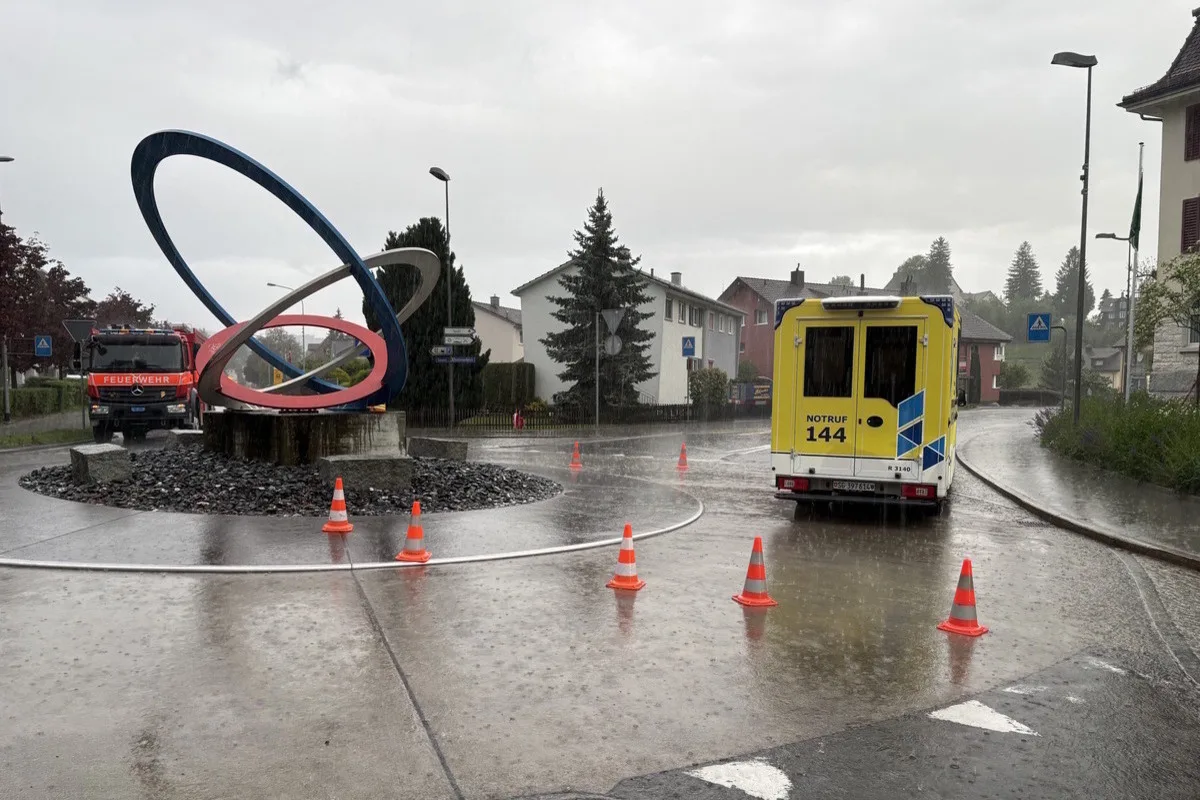 Ein Kreisverkehr mit einem modernen Kunstwerk ist bei regnerischer Witterung zu sehen. Ein Feuerwehrfahrzeug und ein gelbes Ambulanzfahrzeug sind präsent, und Verkehrskegel sind aufgestellt.