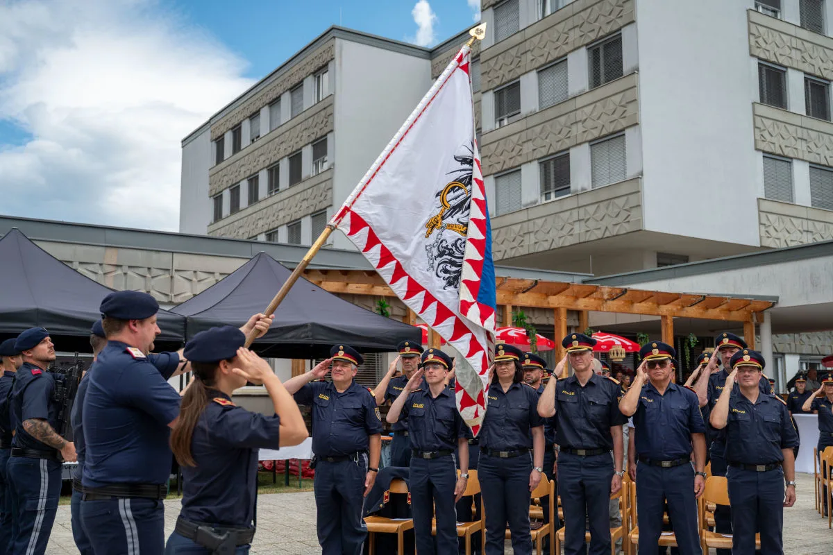 Mehrere Polizisten in Uniform salutieren vor einer gehissten Fahne. Im Hintergrund sind ein Gebäude und Pavillons sichtbar.