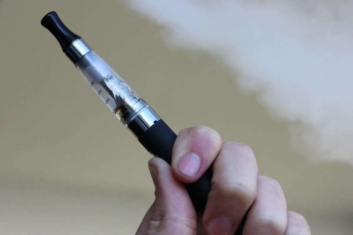 Eine Person hält eine E-Zigarette in der Hand, aus der Dampf aufsteigt. Der Hintergrund ist unscharf.