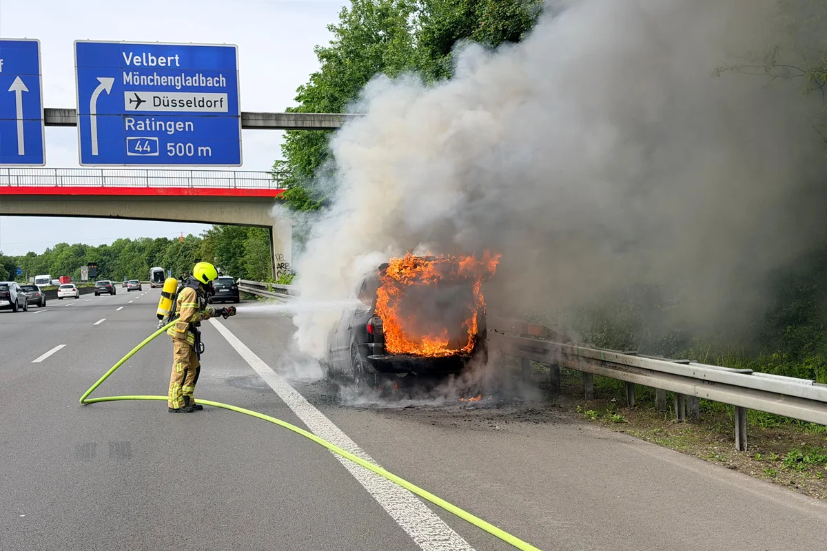Ein brennendes Auto an der Seite der Autobahn, das von einem Feuerwehrmann mit einem Wasserschlauch gelöscht wird. Rauch steigt in die Luft.