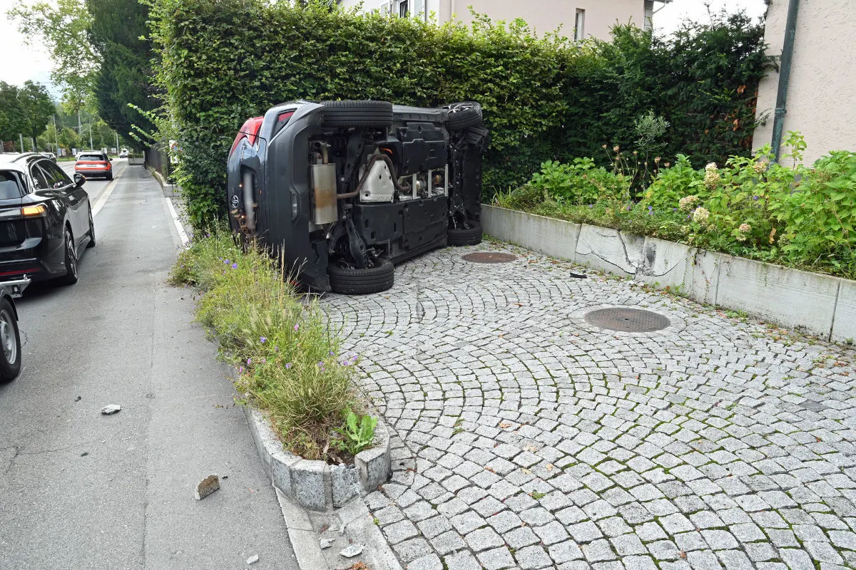 Ein Auto liegt auf der Seite auf einem gepflasterten Bereich neben einer Straße, umgeben von Büschen und Blumen.