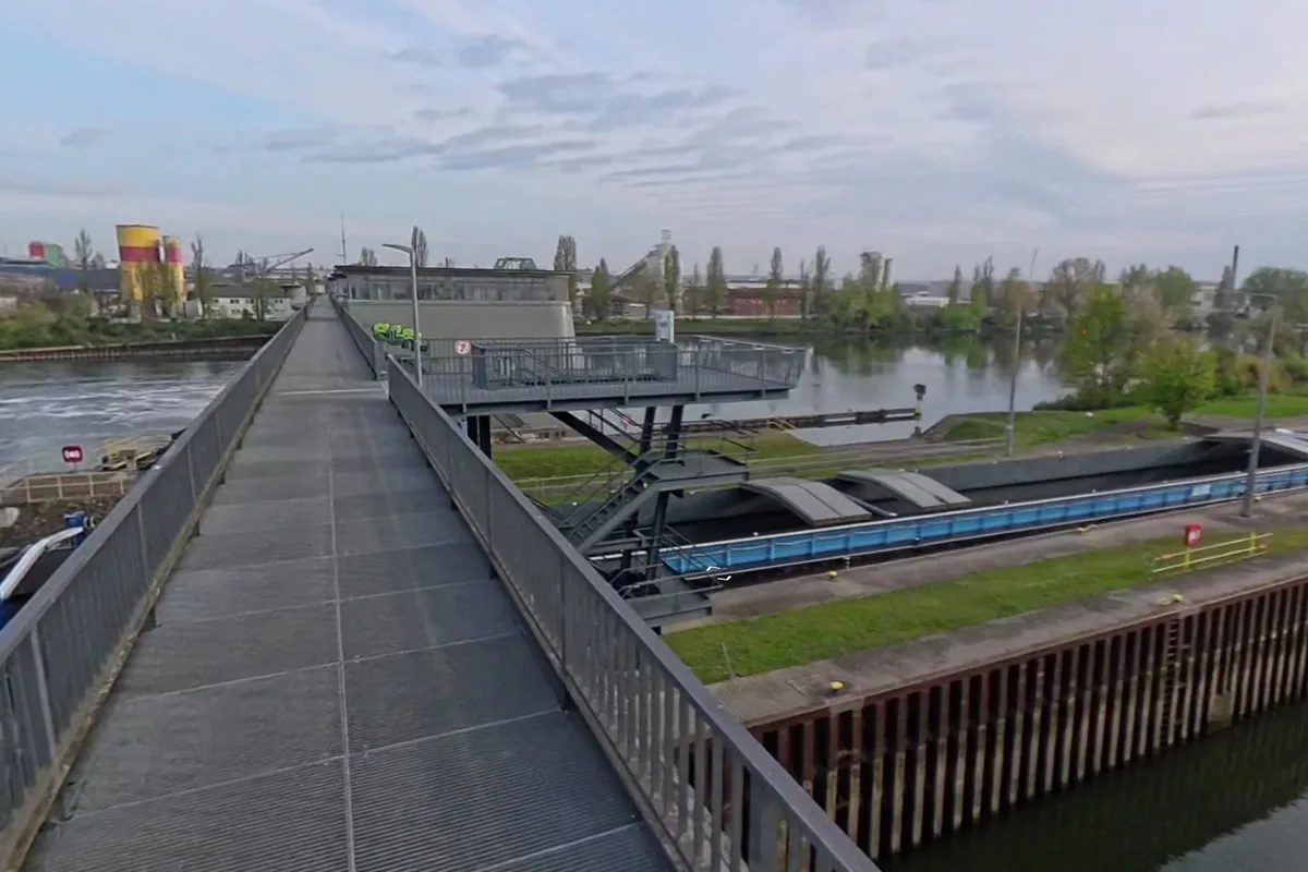 Eine Brücke erstreckt sich über einen Kanal. Im Hintergrund ist eine Industrieanlage sichtbar.