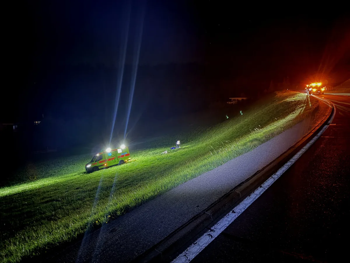 Ein Rettungsfahrzeug mit eingeschaltetem Blaulicht steht auf einer Wiese neben der Autobahn. Einsatzkräfte beleuchten die dunkle Umgebung.