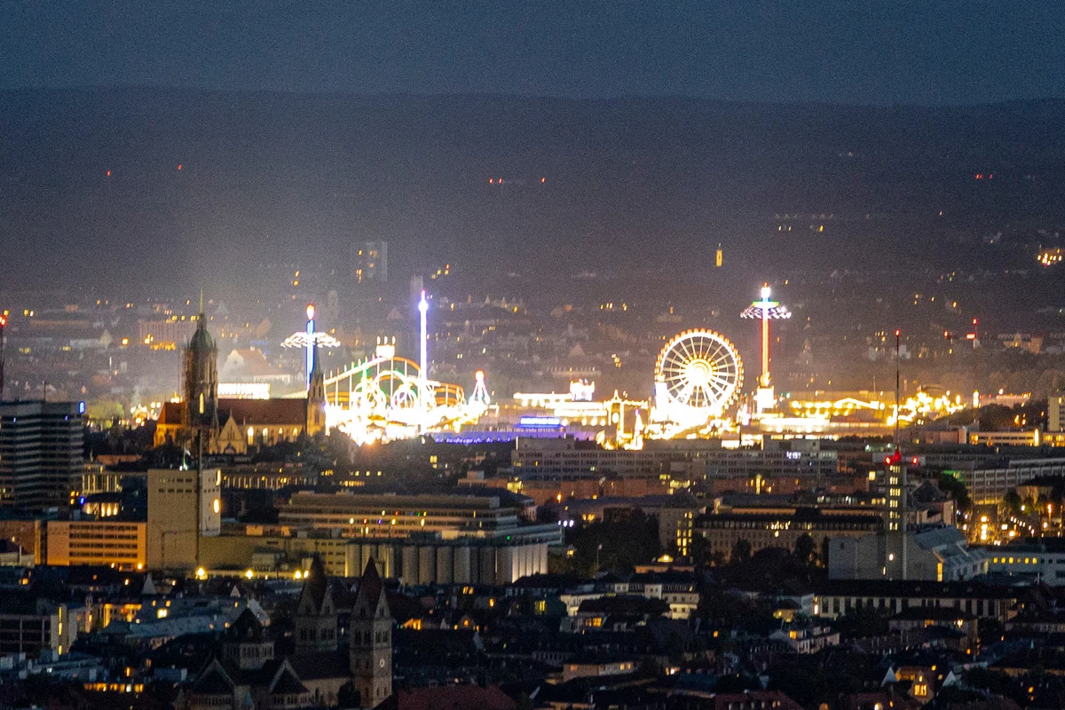 Beleuchtetes Volksfest mit einem großen Riesenrad bei Nacht. Im Hintergrund ist eine Stadtsilhouette sichtbar.