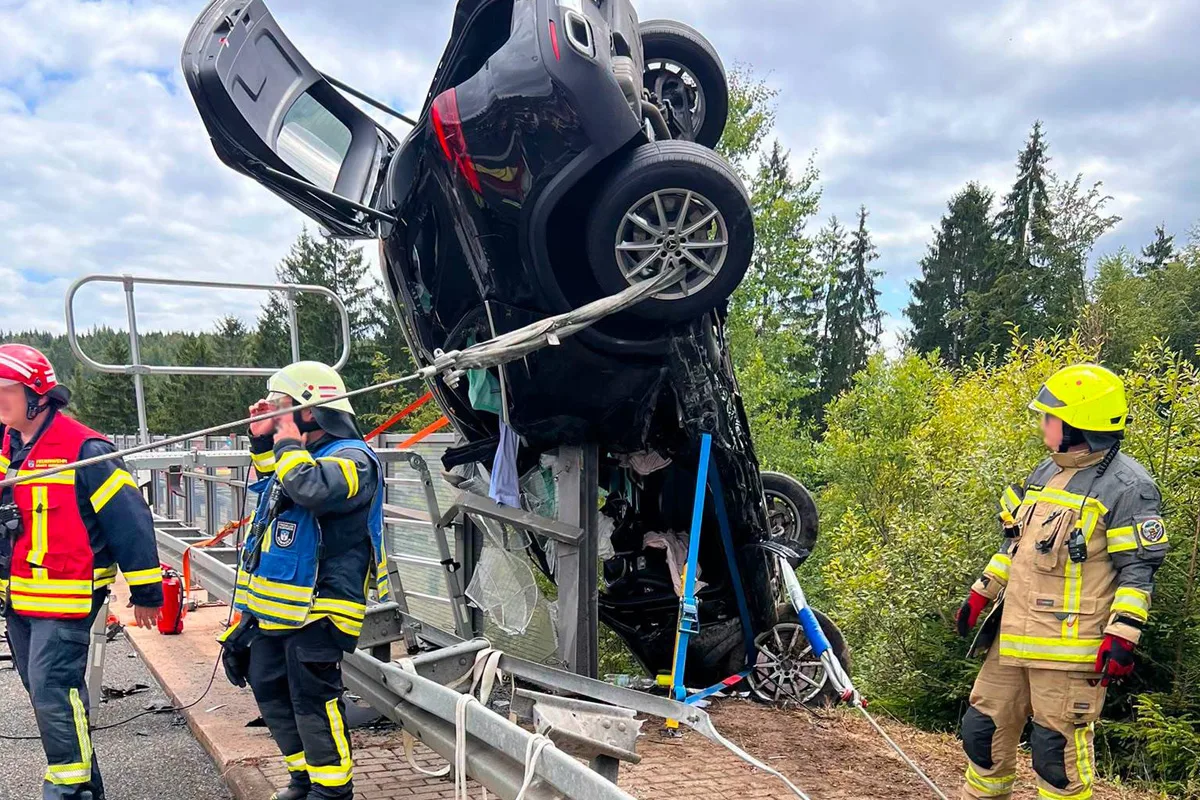 Ein schwarzer Pkw hängt gefährlich an einer Leitplanke über einer Brücke, während Feuerwehrleute ihn sichern. Der Himmel ist bewölkt, und im Hintergrund sind Bäume zu sehen.