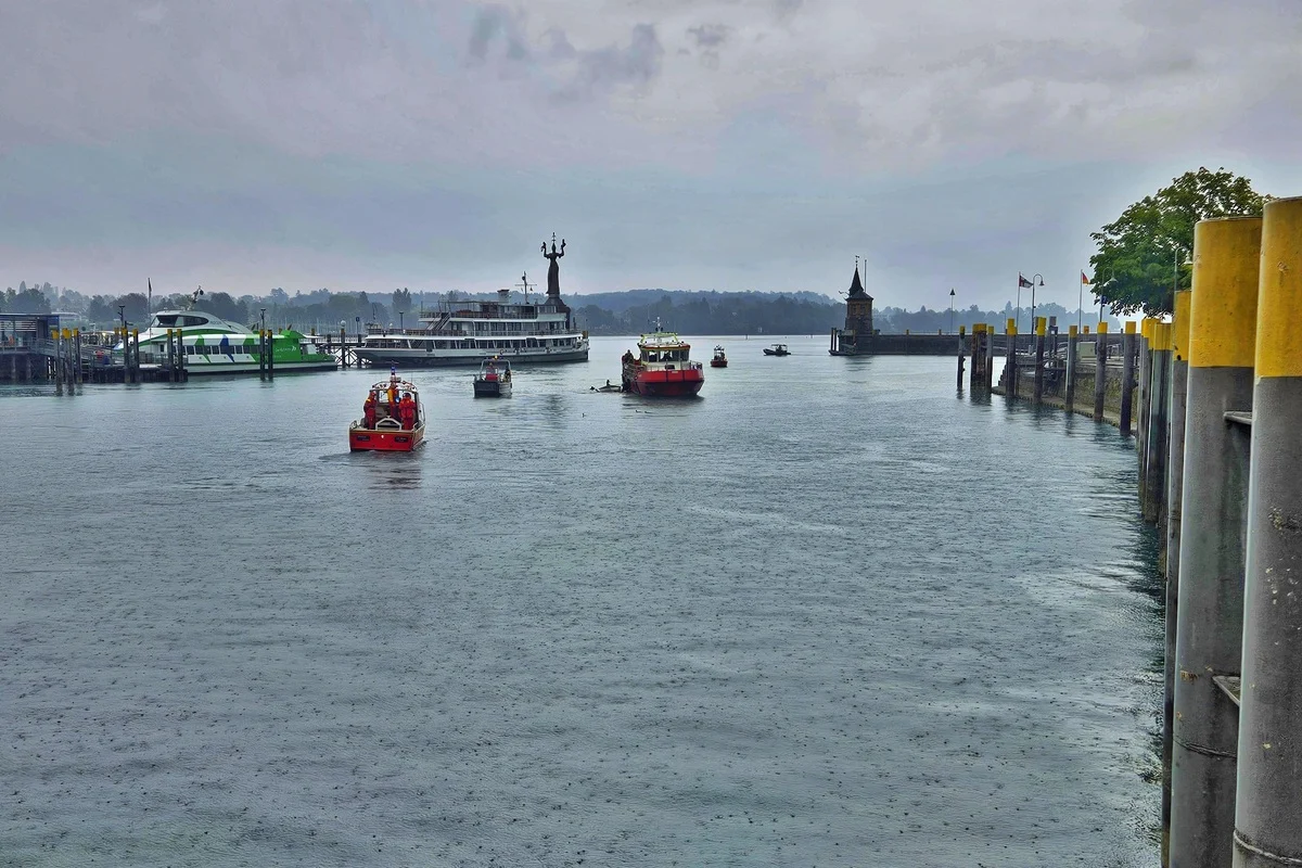 Mehrere Rettungsboote sind bei Regenwetter auf einem großen Gewässer unterwegs. Im Hintergrund sind weitere Boote, eine Hafenanlage und eine Statue zu sehen.