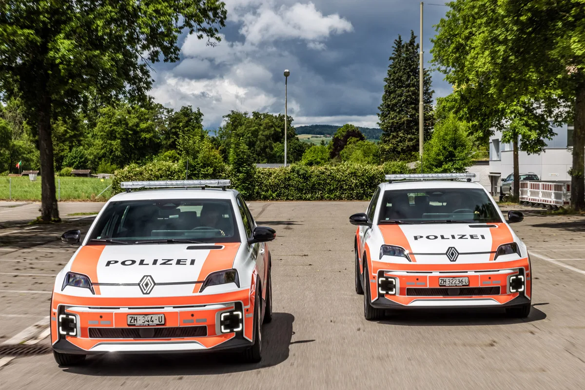 Zwei weiße und orangefarbene Polizeiwagen mit der Aufschrift 'Polizei' fahren nebeneinander auf einer leeren Straße. Im Hintergrund sind Bäume und Himmel zu sehen.